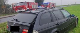 Na łące widać ciemny samochód z uszkodzonym tyłem, w oddali widać trzy samochody straży pożarnej oraz jeden radiowóz Policji