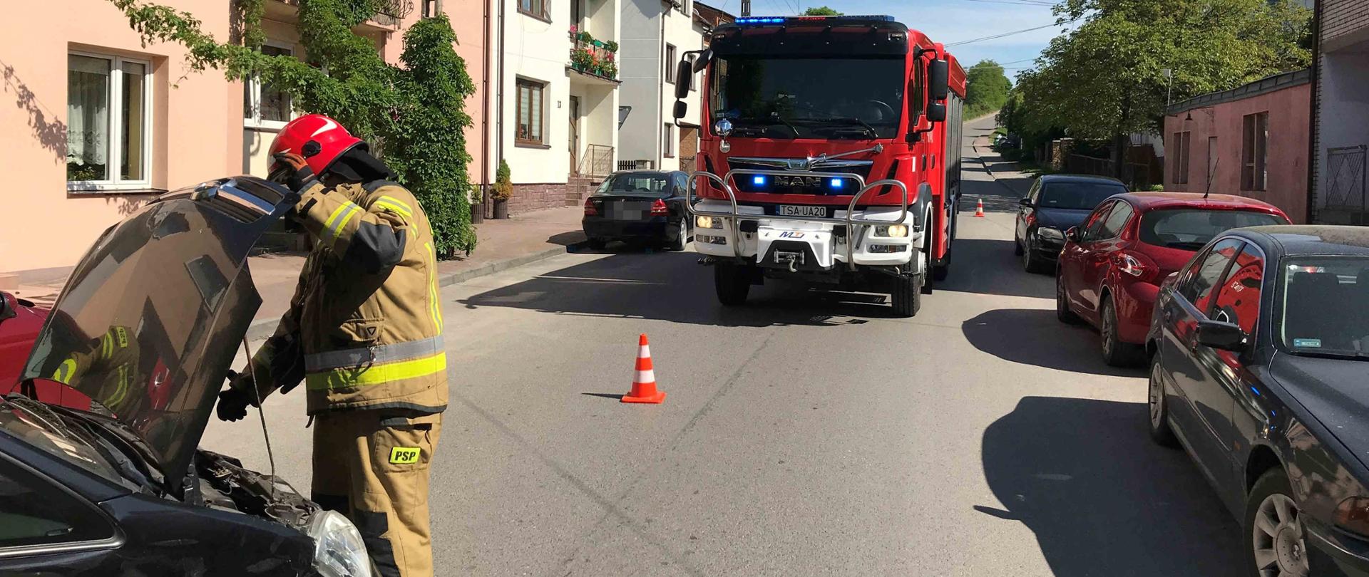 Zdjęcie zrobione na tle budynków mieszkalnych. Na zdjęciu widać jeden rozbity samochód z otwartą maską przy którym stoi strażak. W oddali widać samochód strażacki. Na drodze ustawiony stożek ostrzegawczy, wzdłuż drogi zaparkowane samochody osobowe.
