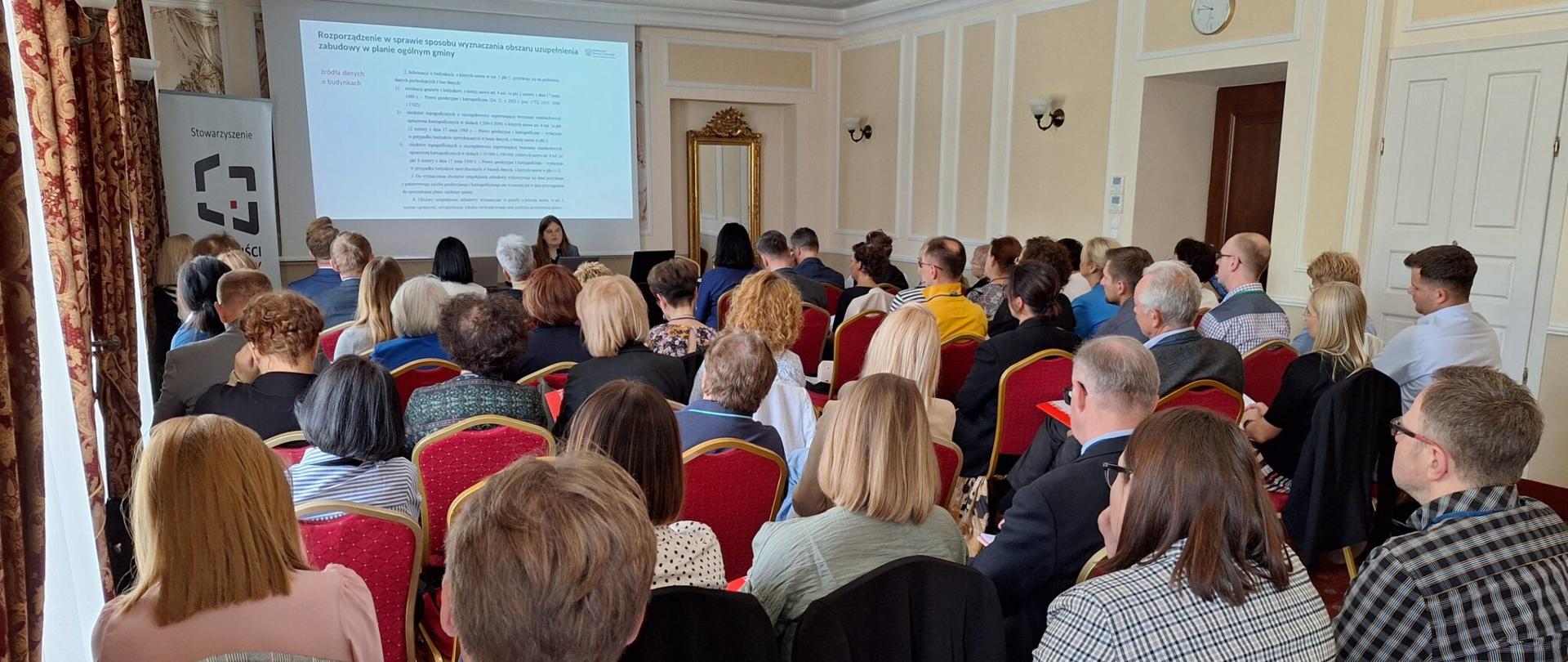 Sala konferencyjna w beżowo-białych odcieniach. Na pierwszym planie widać ludzi siedzących na krzesłach. W oddali siedzi kobieta, za którą wyświetlana jest prezentacja. Z lewej strony stoi baner reklamowy, po prawej stronie wisi lustro.