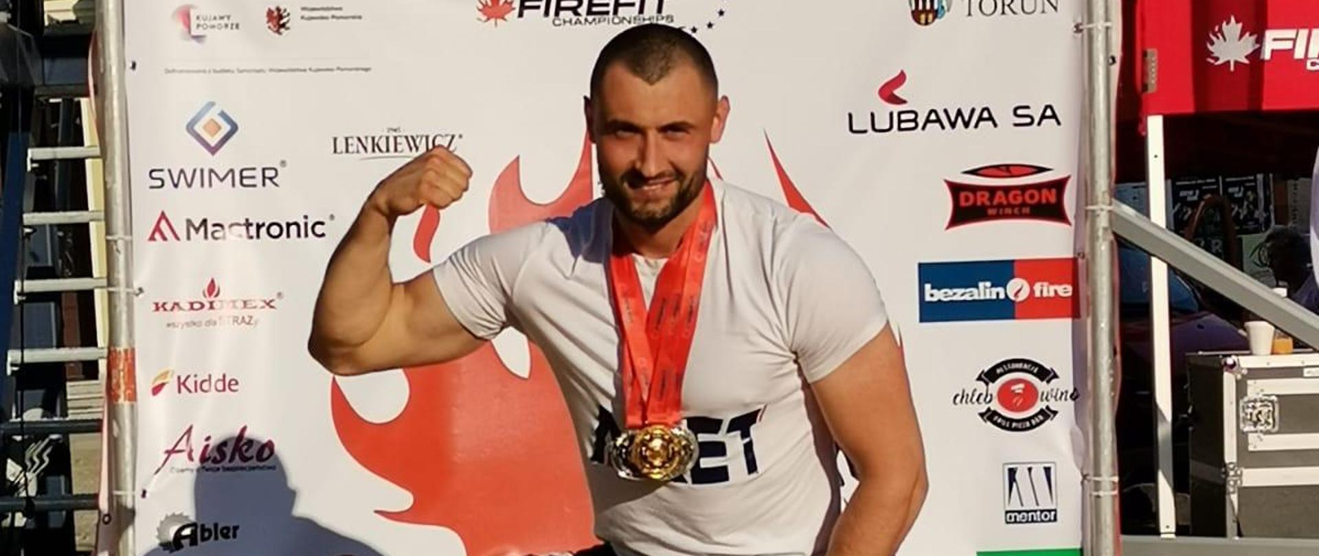 mł. kpt. Adam Spychał na podium z medalami i nagrodami (toporek na desce). W tle ścianka zawodów ze sponsorami.