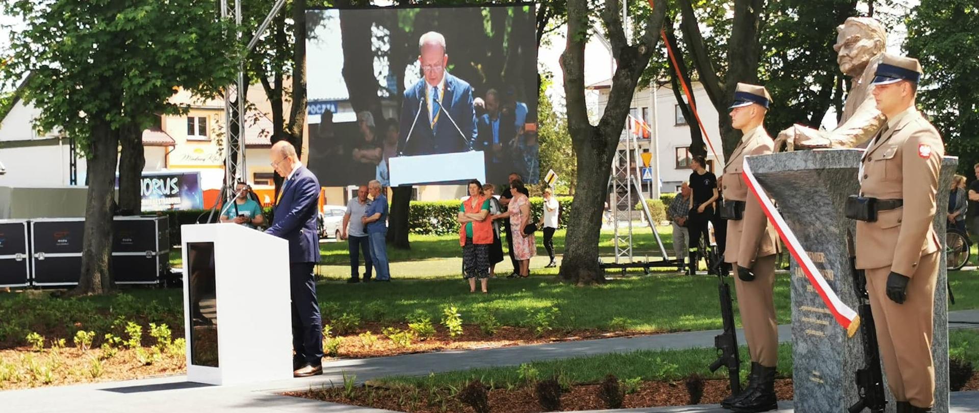 Na pierwszym planie znajduje się pomnik Premiera Jana Olszewskiego, przy którym stoi warta honorowa Wojska Polskiego. Po lewej stronie zdjęcia, za mównicą stoi ubrany w granatowy garnitur mężczyzna, który odczytuje list. W tle znajduje się telebim. 