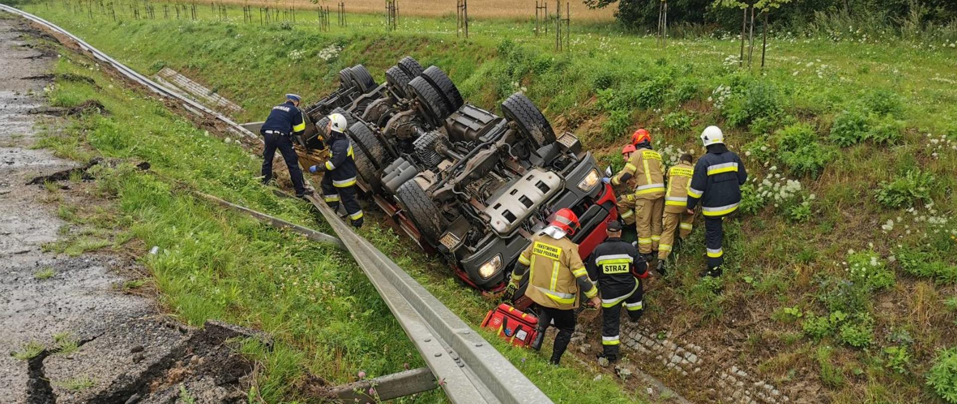 Przewrócony samochód ciężarowy w przydrożnym rowie. Przy kabinie strażacy w trakcie działań.