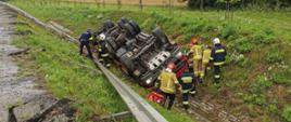 Przewrócony samochód ciężarowy w przydrożnym rowie. Przy kabinie strażacy w trakcie działań.