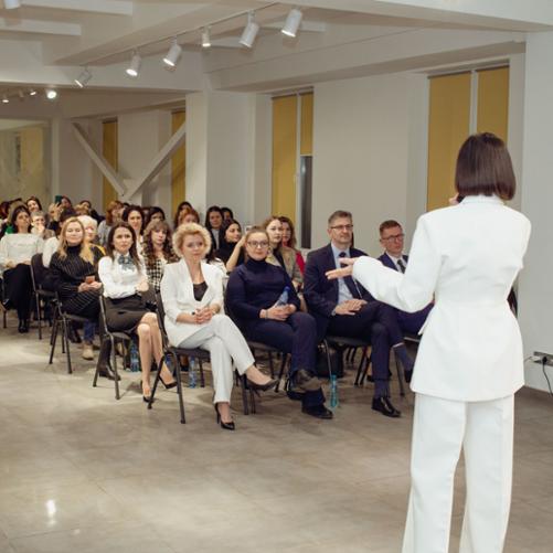 Sala konferencyjna. Kobiety siedzą na krzesłach ustawionych w rzędach. Jedna kobieta stoi przed nimi i przemawia do mikrofonu. 