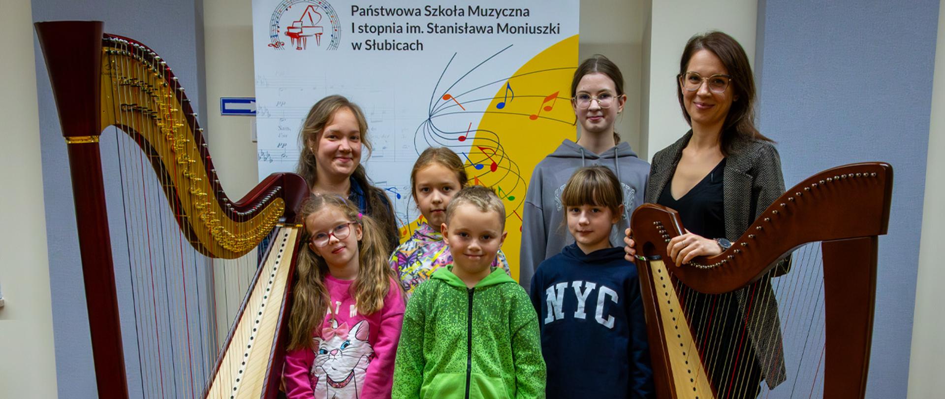 Dwie nauczycielki i pięcioro uczniów stoją przy dwóch harfach.