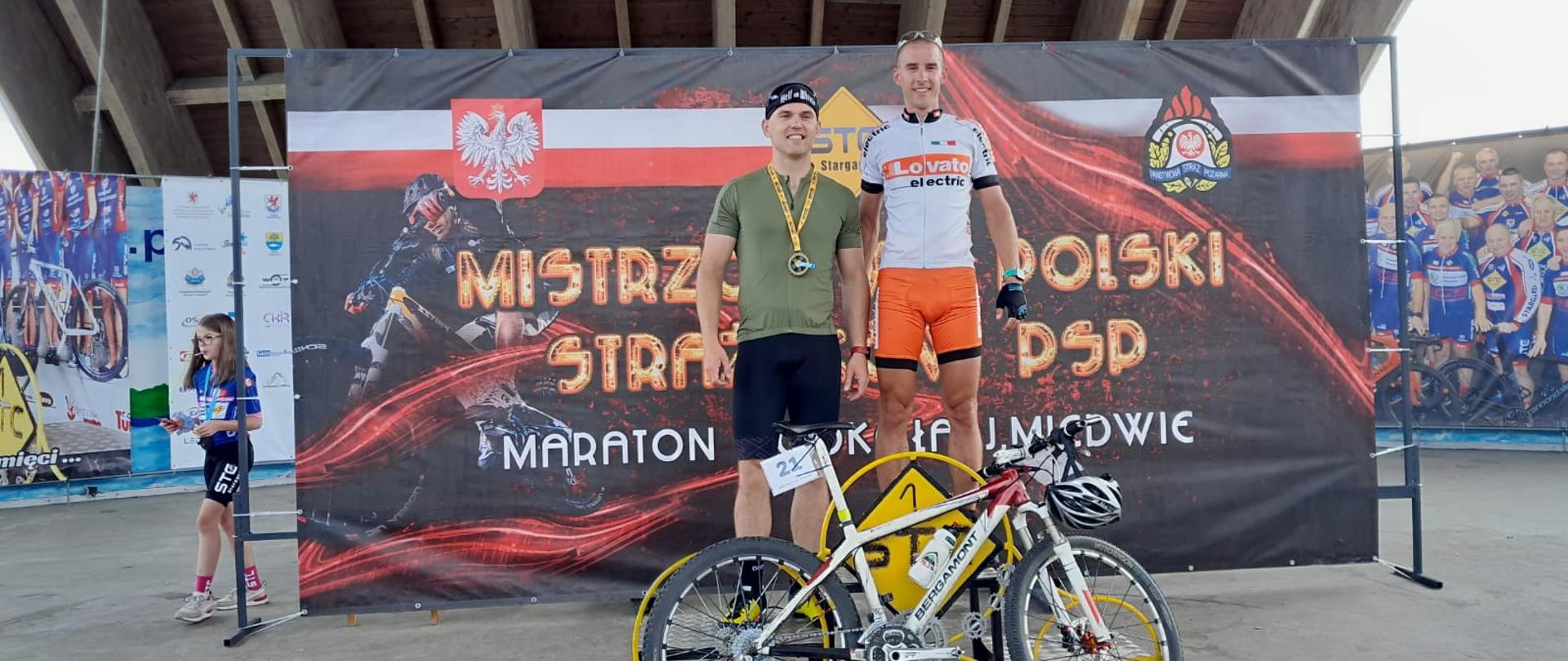 Dwóch zawodników w sportowym w stroju sportowym, stojący na podium, pozujący za rowerem, na tle banneru z napisem "Mistrzostwa Polski Strażaków w PSP"