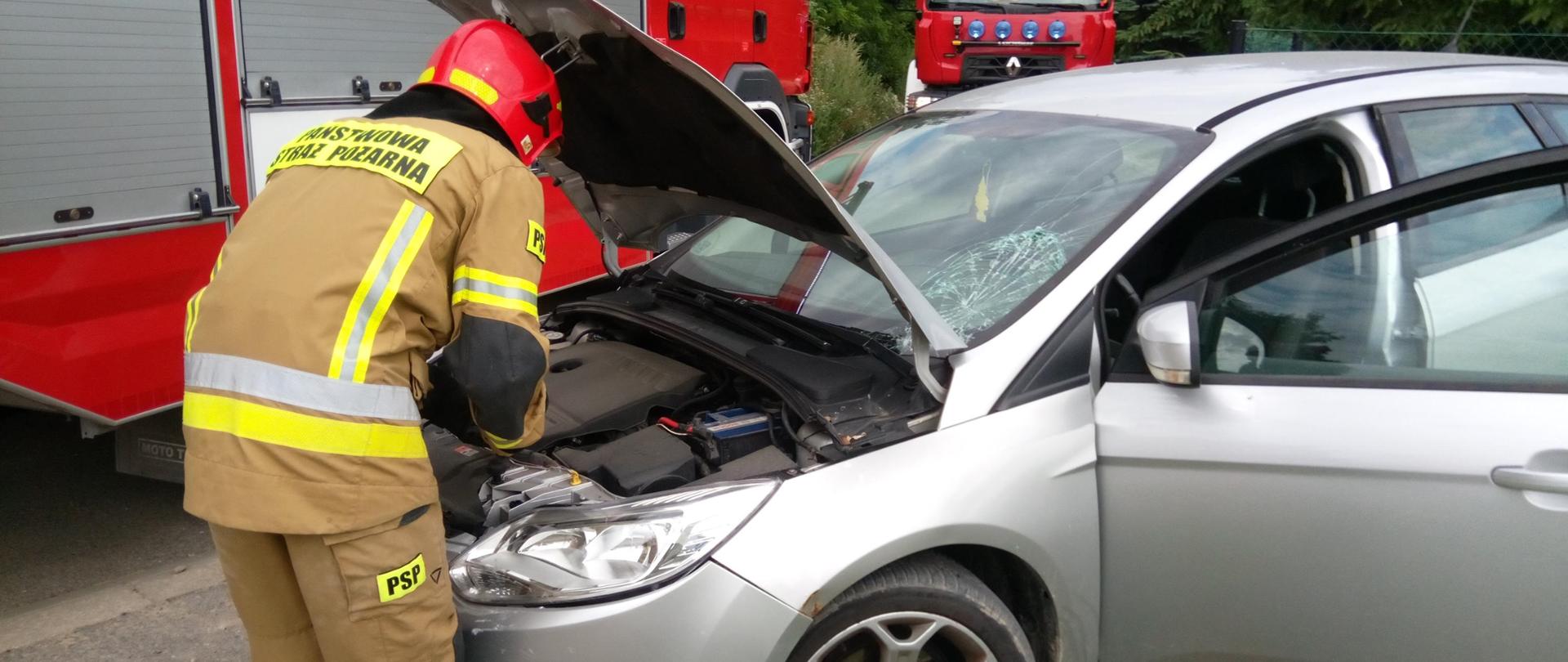 Umundurowany strażak odkręca akumulator w srebrnym samochodzie osobowym.