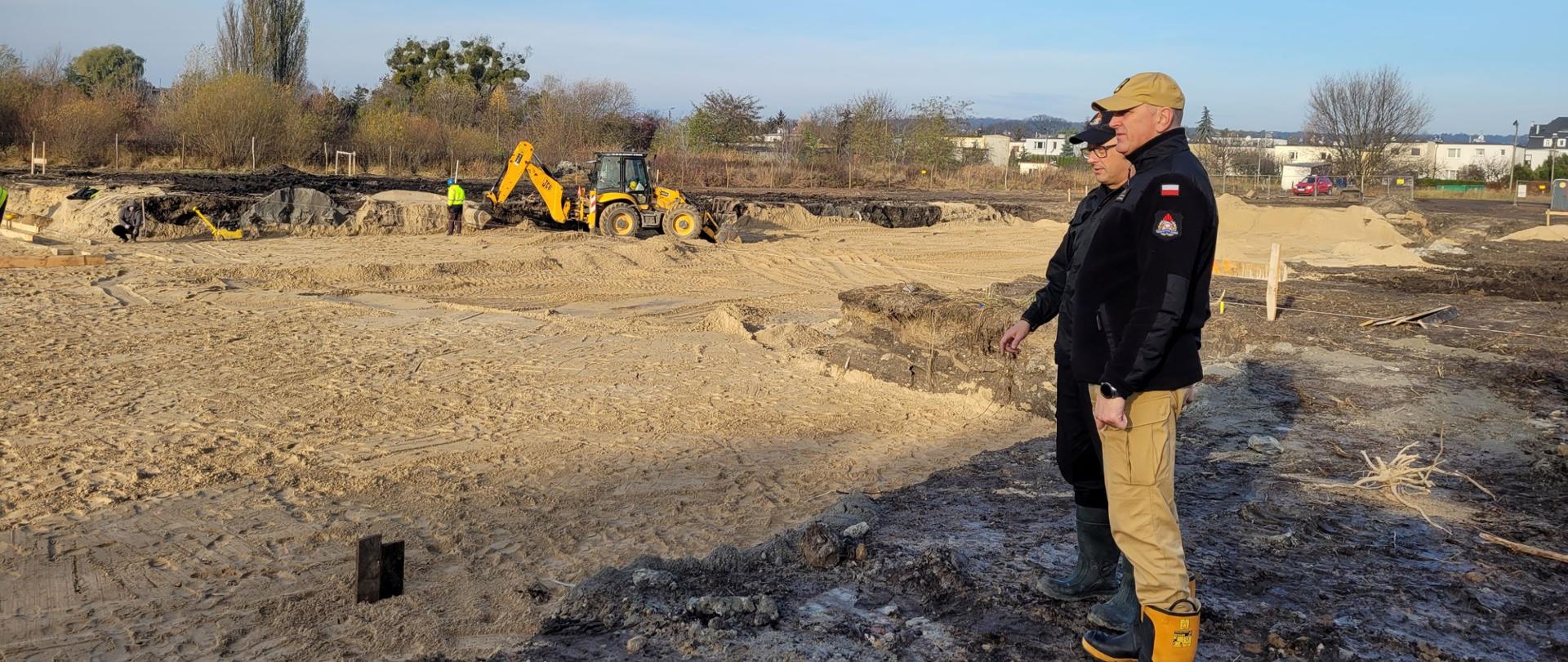 Zdjęcie przedstawia dwóch strażaków stojących nad wykopem na placu budowy. Wewnątrz wykopu trwają prace ziemne wykonywane przez ludzi i żółtą koparkę . W tle drzewa, krzewy i budynki.