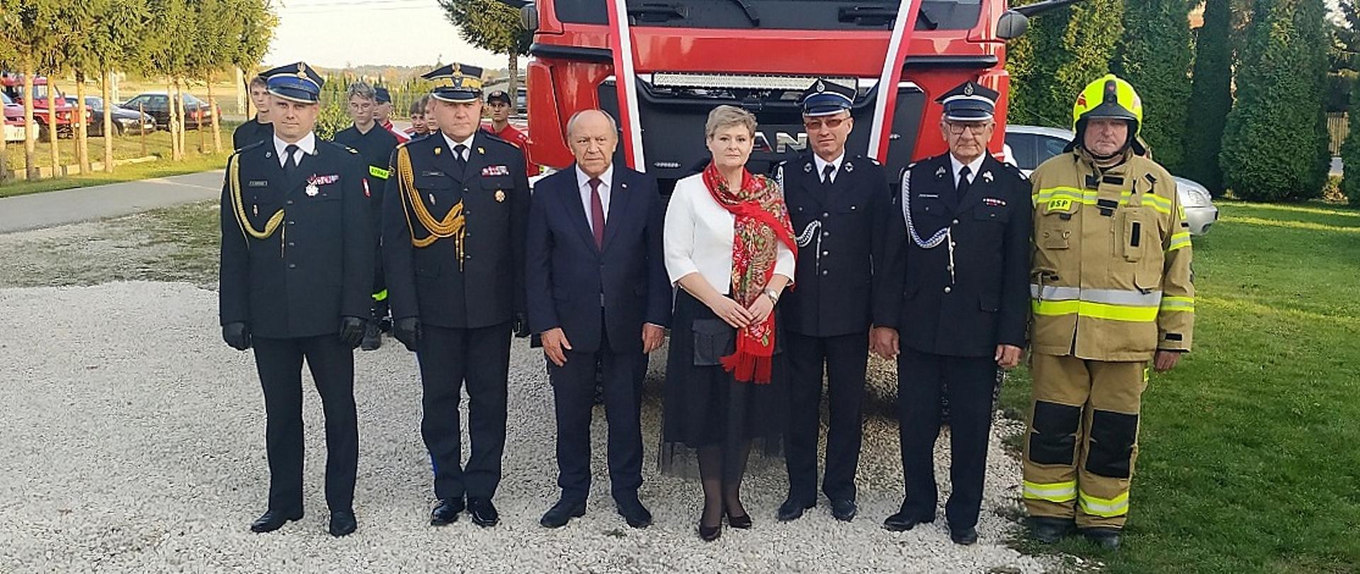 Fotografia grupowa upamiętniających gości na tle nowo przekazanego samochody ratowniczo-gaśniczego. Od prawej stoją ubrani w mundury galowe koloru granatowego Państwowej Straży Pożarnej: komendant powiatowy oraz podkarpacki komendant wojewódzki, następnie w środku: poseł na sejm RP oraz burmistrz miasta i gminy Przecław, a po lewej stronie druhowie jednostki OSP Tuszyma ubrani w galowe mundury koloru granatowego i ubranie bojowe typu nomex koloru piaskowego. 