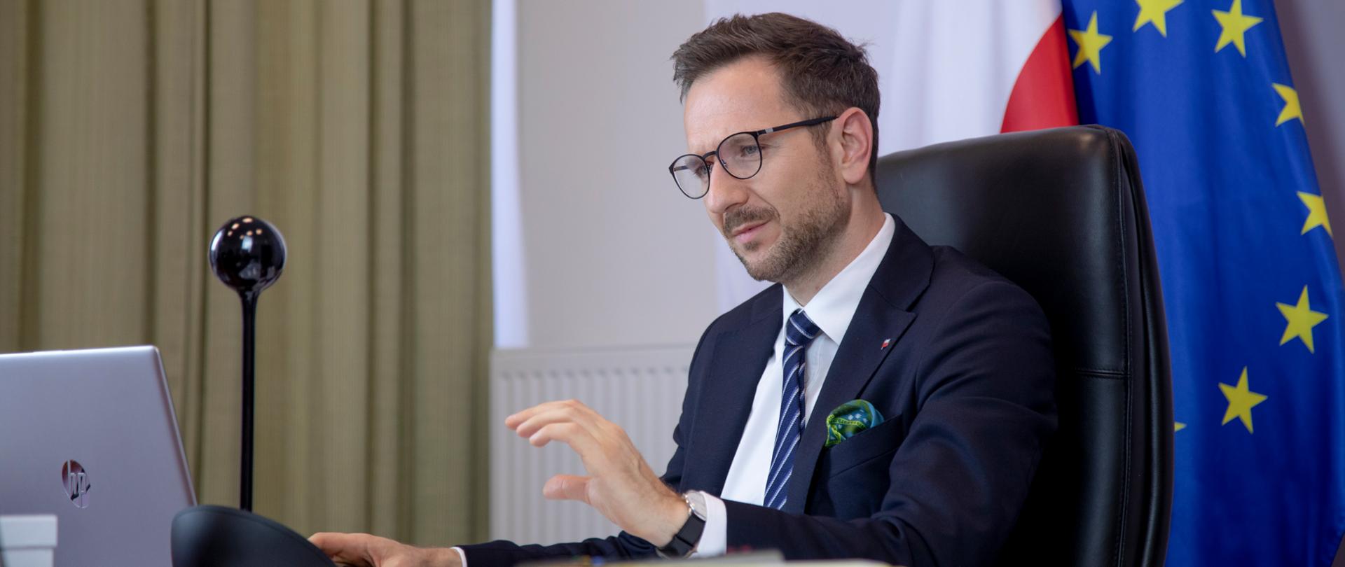 wiceminister Waldemar Buda przed komputerem uczestniczy w spotkaniu online, za nim flagi Polski i Unii Europejskiej
