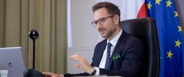 wiceminister Waldemar Buda przed komputerem uczestniczy w spotkaniu online, za nim flagi Polski i Unii Europejskiej