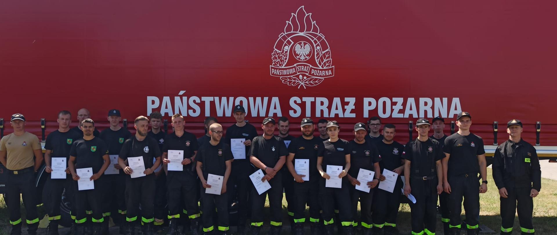 Zdjęcie grupowe kursantów z zaświadczeniami na tle naczepy strażackiej z logiem i napisem Państwowa Straż Pożarna.
