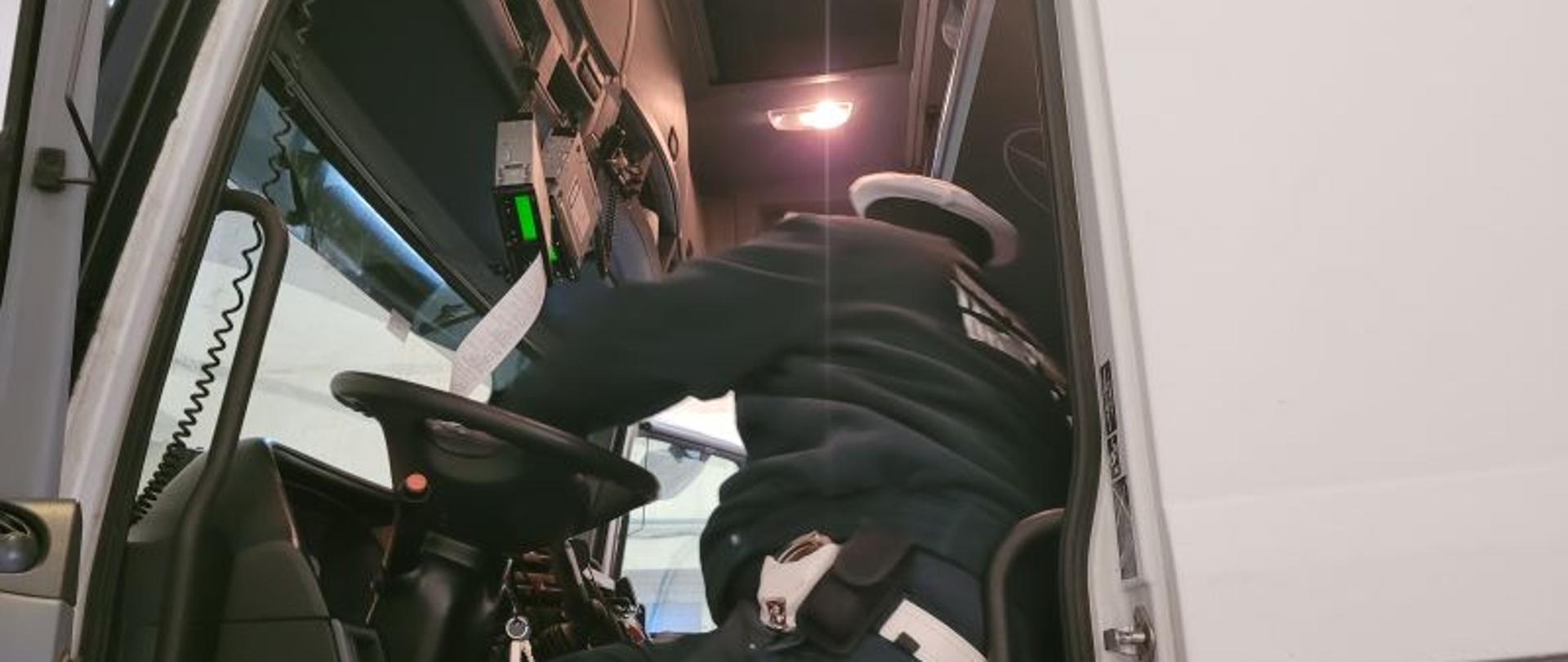 Umundurowany funkcjonariusz kujawsko-pomorskiej Inspekcji Transportu Drogowego siedzi na fotelu kierowcy w kabinie ciężarówki. Inspektor kontroluje prawidłowość działania tachografu oraz instalację urządzenia rejestrującego czasy pracy kierowcy.