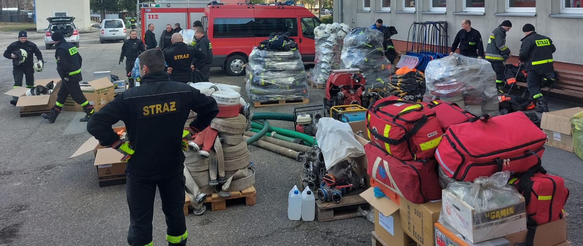 Na zdjęciu widoczny jest sprzęt składowany na placu wewnętrznym Komendy Miejskiej PSP w Tarnowie. Na pierwszym planie widoczne są zestawy PSP R-1 oraz węże pożarnicze, a strażacy kompletują ten sprzęt.