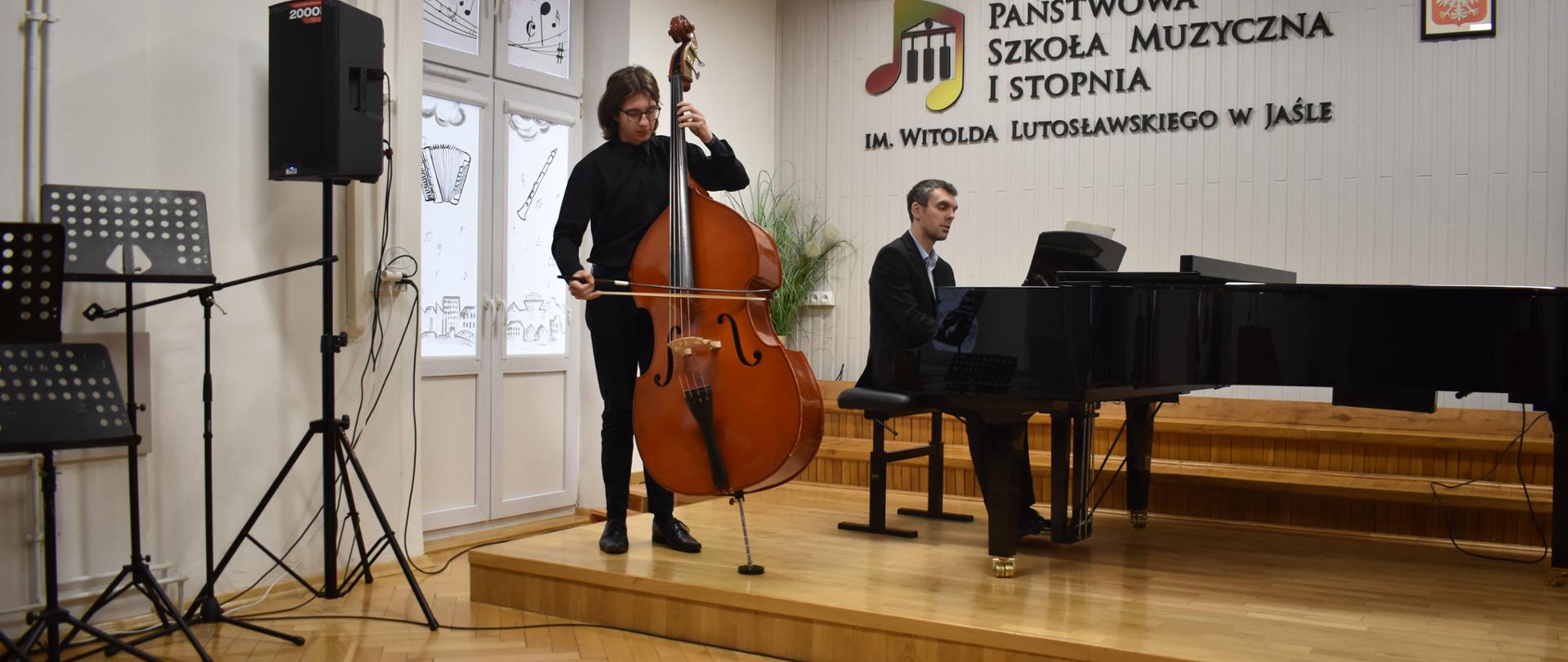 Na zdjęciu wykonanym w auli PSM widnieje uczeń grający na kontrabasie z akompaniamentem fortepianu. Przy fortepianie nauczyciel Mateusz Gałuszka. Kolorystyka zdjęcia jest biało-brązowa.