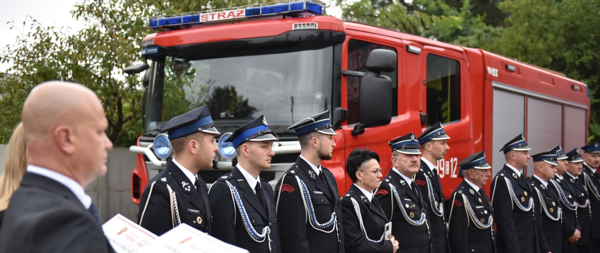 strażacy osp stojący w szeregu, za nimi stoi samochód strażacki, po lewej mężczyzna trzymający białe tabliczki obraz rozmazany