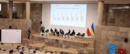 Aula sali z uczestnikami obrad XV Międzynarodowego Forum „Polsko-Ukraińskie dni biznesowe”, w centralnej części fotografii podest z uczestnikami panelu, którzy patrzą na prezentację na telebimie.
