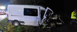 Kolizja na drodze wojewódzkiej. Zdjęcie przedstawia samochód typu bus widziany z prawego boku. Pojazd ma bardzo mocno uszkodzony przód. Z lewej strony widać fragmenty wozów strażackich z włączonymi sygnałami błyskowymi koloru niebieskiego. Na jezdni stoja pachołki ostrzegawcze. 