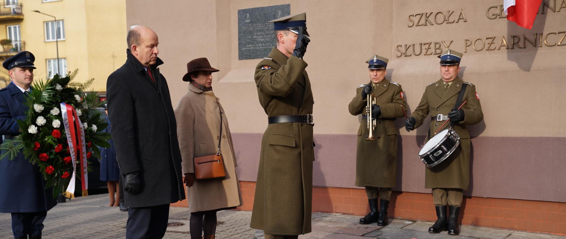 Zdjęcie przedstawia moment składania wieńców przed tablicą upamiętniającą strajk w Wyższej Oficerskiej Szkole Pożarniczej, która znajduje się przy Szkole Głównej Służby Pożarniczej w Warszawie. Na pierwszym planie zdjęcia znajduje się żołnierz Wojska Polskiego, asystujący w ceremonii składania wieńców. Tuż za nim znajdują się elegancko ubrani kobieta i mężczyzna. Na drugim planie zdjęcia znajdują się pozostali uczestnicy wydarzenia. 