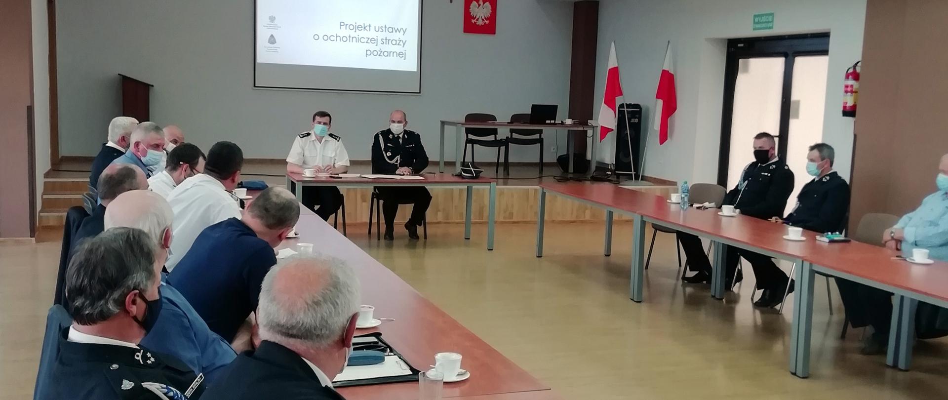 Zdjęcie zrobione w sali szkoleniowej. Przy stołach siedzą mężczyźni. Część z nich ubrana jest w mundury osp. Na ekranie widać stronę prezentacji z napisem: projekt ustawy o ochotniczej straży pożarnej