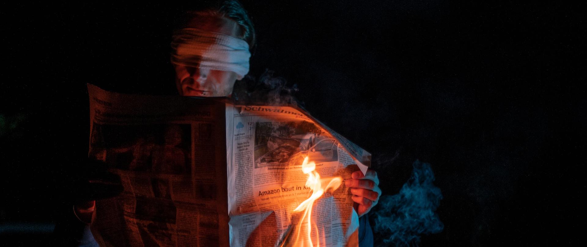 Mężczyzna z opaską na oczach trzymający płonącą gazetę.