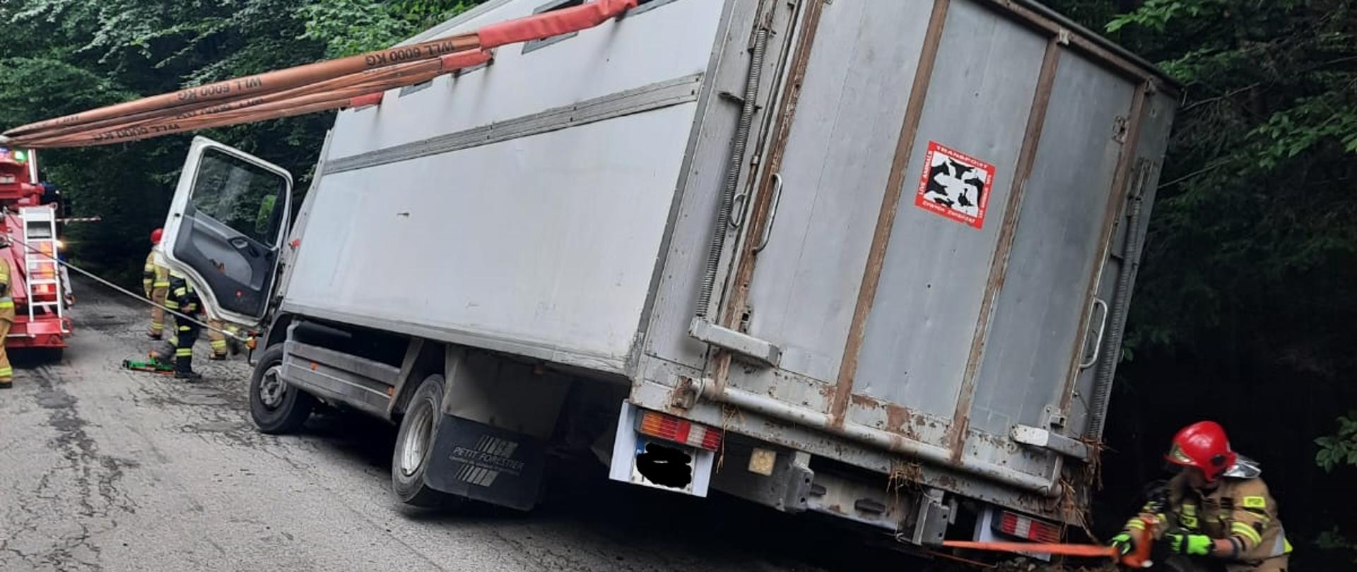Na zdjęciu widać samochód ciężarowy przeznaczony do transportu bydła, który zjechał na grząskie pobocze jezdni i mocno się przechylił. Pojazd jest zabezpieczony za pomocą lin przed przewróceniem się na bok. Przy pojeździe widać samochód strażacki i strażaków. 