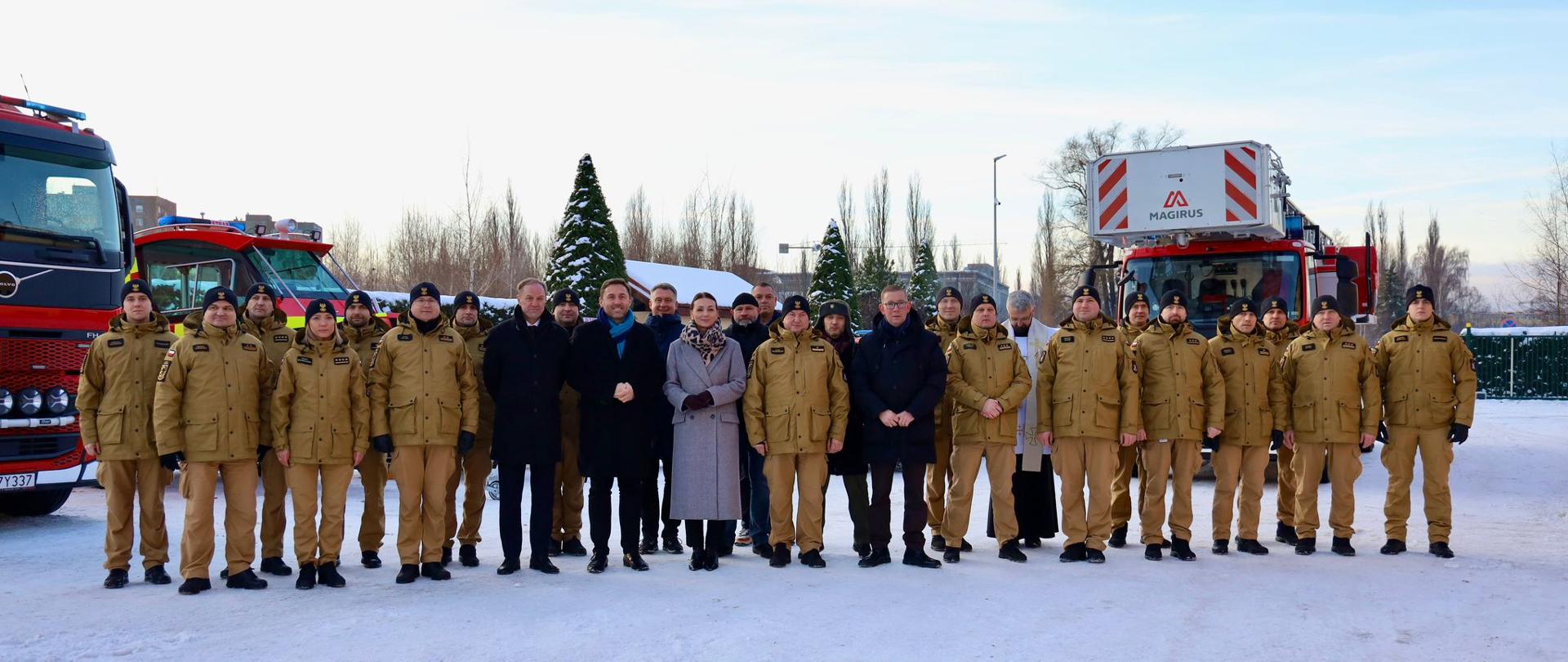 Grupa osób, głownie strażaków, stoi na placu przed wozami pożarniczymi