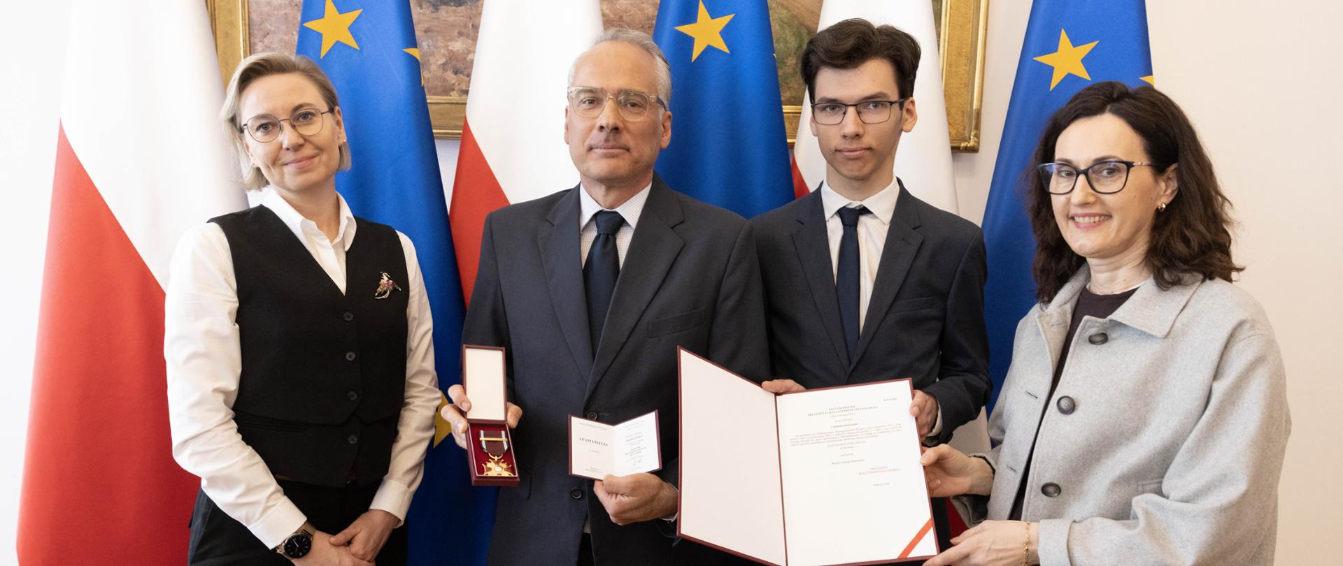 Ministra do spraw Społeczeństwa Obywatelskiego Adriana Porowska przekazała rodzinie pani doktor Moniki Madalińskiej, nadany jej pośmiertnie Złoty Krzyż zasługi. W tle flagi Polski i Unii Europejskiej.