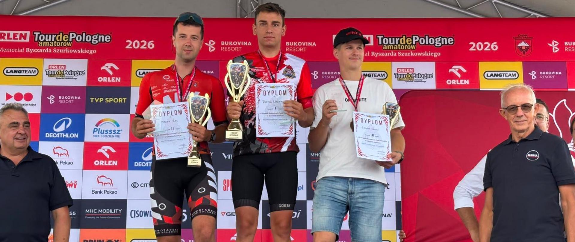 Troje kolarzy na podium wyścigu Tour de Pologne Amatorów 2026. Zwycięzcy trzymają puchary i dyplomy w asyście dwóch mężczyzn po bokach podium.