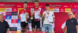 Troje kolarzy na podium wyścigu Tour de Pologne Amatorów 2026. Zwycięzcy trzymają puchary i dyplomy w asyście dwóch mężczyzn po bokach podium.