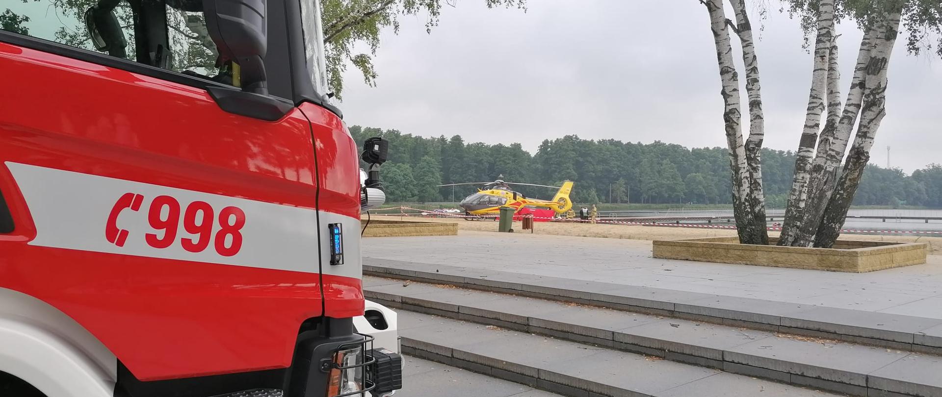 Z lewej strony przód kabiny pojazdu pożarniczego. Na pierwszym planie schody z kostki brukowej. W tle widoczny helikopter LPR stojący na plaży, jeziora i las.