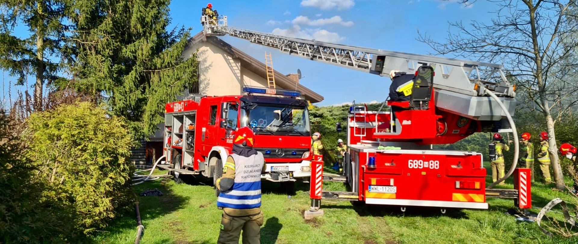Na zdjęciu widać dwa czerwone wozy strażackie. Wóz z prawej strony jest z drabiną z koszem. W koszu są dwaj strażacy i zbliżają się do budynku. Za pojazdami stoi budynek mieszkalny, elewacja koloru jasnego, dach koloru brązowego. Spod dachu wydobywa się jasny dym. Przed pojazdami na trawie stoi umundurowany strażak w czerwonym hełmie i kamizelce z napisem „Kierujący działaniem ratowniczym”.