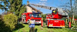 Na zdjęciu widać dwa czerwone wozy strażackie. Wóz z prawej strony jest z drabiną z koszem. W koszu są dwaj strażacy i zbliżają się do budynku. Za pojazdami stoi budynek mieszkalny, elewacja koloru jasnego, dach koloru brązowego. Spod dachu wydobywa się jasny dym. Przed pojazdami na trawie stoi umundurowany strażak w czerwonym hełmie i kamizelce z napisem „Kierujący działaniem ratowniczym”.