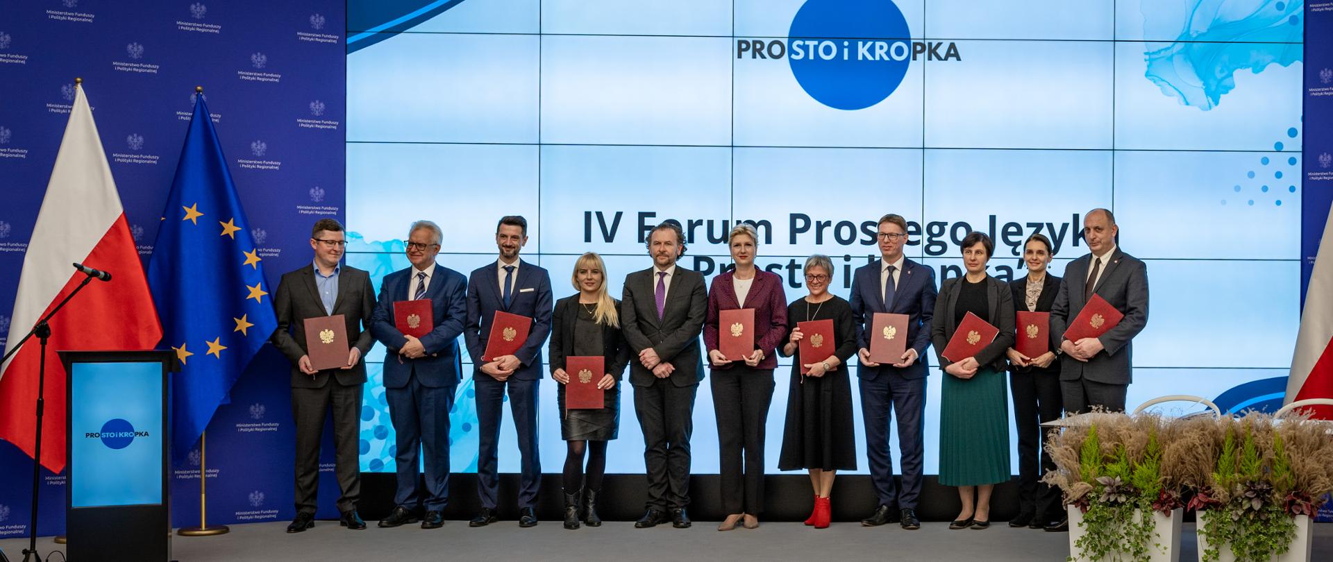 Na scenie w sali konferencyjnej grupa osób stoi w szeregu. Za nimi na ekranie napis: "Prosto i kropka". Obok flagi PL i UE, kwiaty w donicach i mównica.