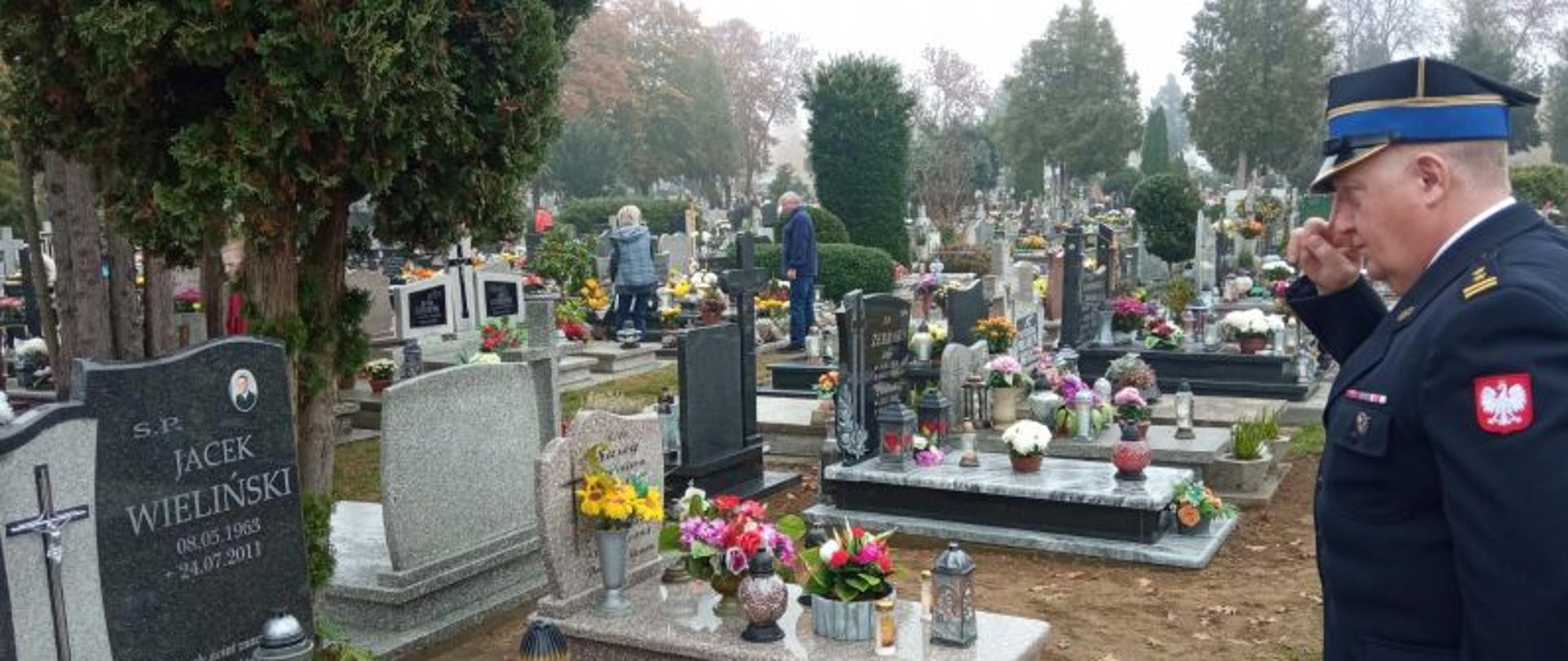 Zdjęcie wykonane na cmentarzu. Po lewej stronie salutujący strażak w galowym mundurze z czapką. Na grobach dużo zniczy, Dzień jest pochmurny, jest szaro.