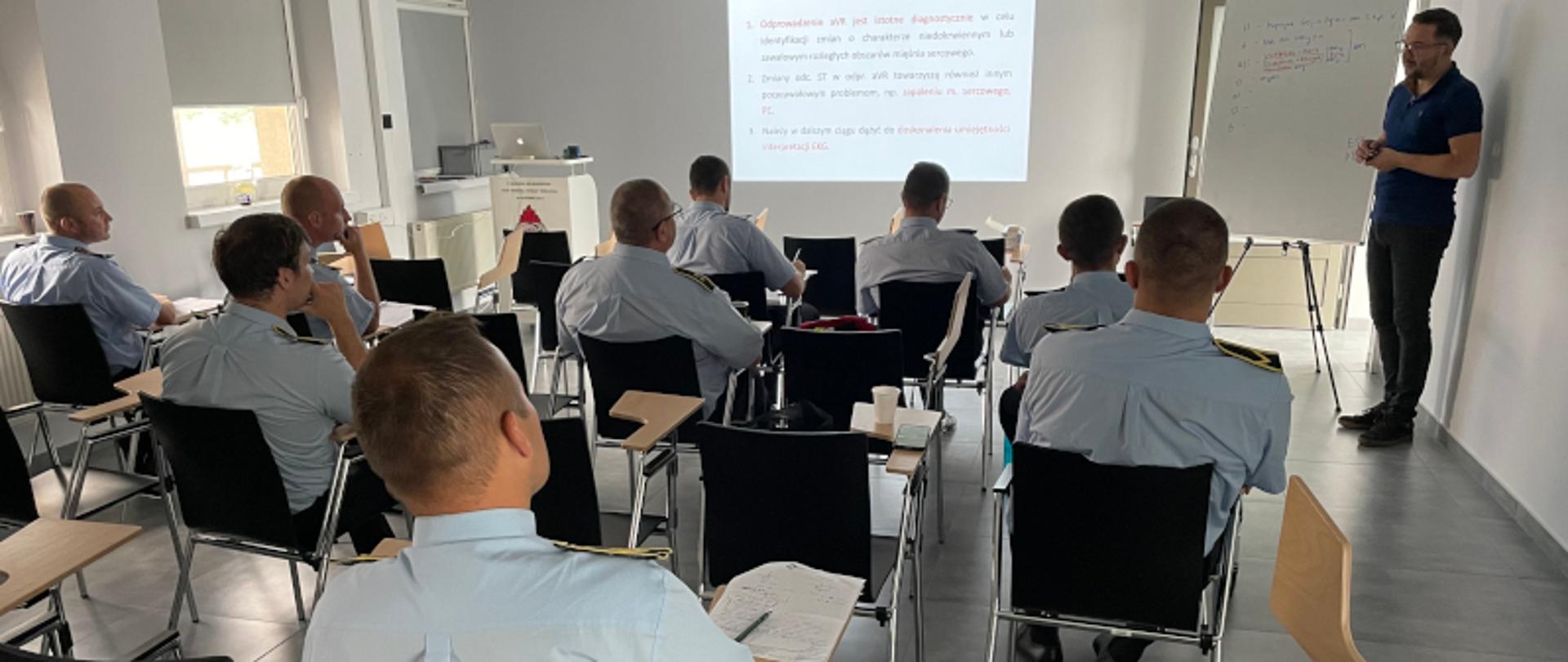 W sali szkoleniowej odwróceni plecami uczestnicy spotkania siedzą na krzesłach, przy tablicy stoi moderator spotkania, na ścianie wyświetla się obraz z rzutnika.