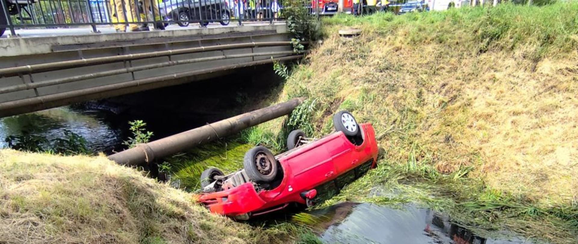 Na zdjęciu czerwony samochód leży do góry kołami w korycie rzeki. Auto częściowo zanurzone jest w wodzie, a koła skierowane są do góry. Po lewej stronie widać strome skarpy porośnięte trawą. W tle znajduje się most z barierką, za którym stoją służby ratunkowe. Widać, że jest to poważny wypadek drogowy.