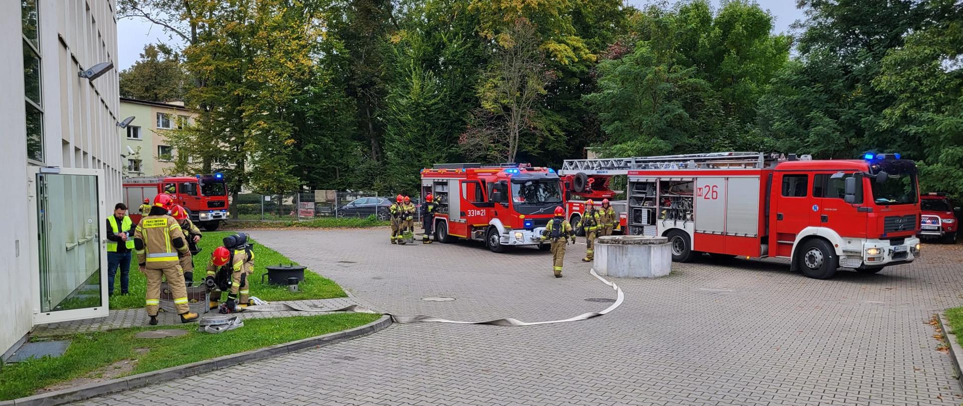 Zdjęcie wykonane przed jednym z budynków szpitala. Na zdjęciu widać ustawione pojazdy pożarnicze. Pojazdy w kolorze czerwonym ze srebrnymi i białymi elementami zabudowy. Przed pojazdami widać strażaków ubranych w ubrania specjalne w kolorze piaskowym i aparaty powietrzne na plecach. Strażacy rozwijają linie gaśnicze z węży pożarniczych podczas ćwiczeń. 