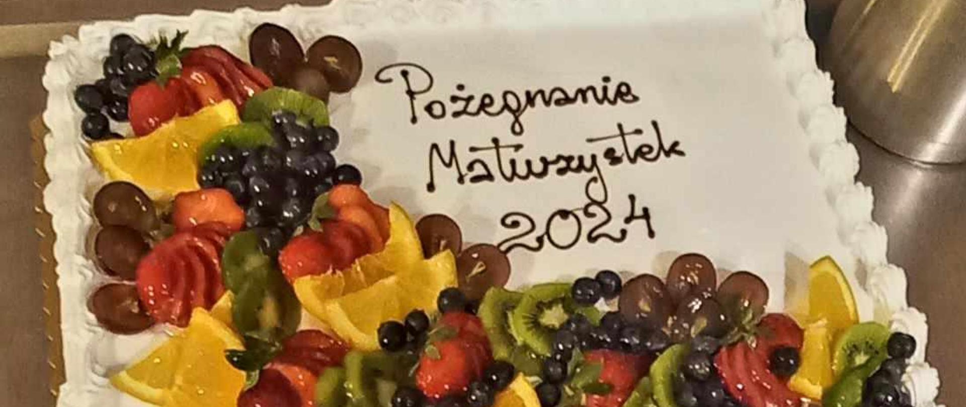 Tort w kształcie kwadrata, z napisem: Pożegnanie Maturzystek 2024. Na torcie różnorodne owoce.