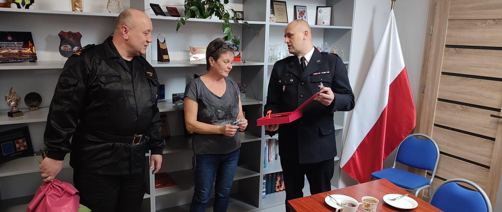 Na zdjęciu widać zastępcę oraz komendanta powiatowego PSP w Ustrzykach Dolnych którzy trzymają w rękach upominki. Między nimi stoi Pani Barbara. Za nimi wszystkimi znajdują się półki na których stoją różne rzeczy. Przed nimi widać biurko na którym są ciasta i kawa. Komendant ubrany jest w mundur galowy, jego zastępca natomiast w ubranie dowódczo-sztabowe. 