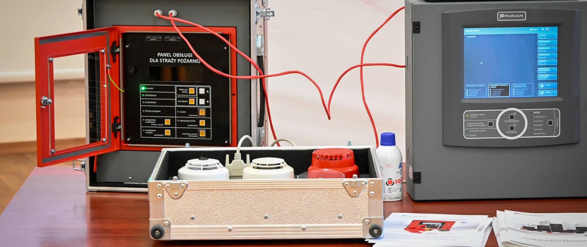 Zbliżenie na urządzenia demonstracyjne systemu sygnalizacji pożarowej: panel obsługi w walizce, centralę z ekranem dotykowym oraz czujki dymu umieszczone w aluminiowej skrzynce.