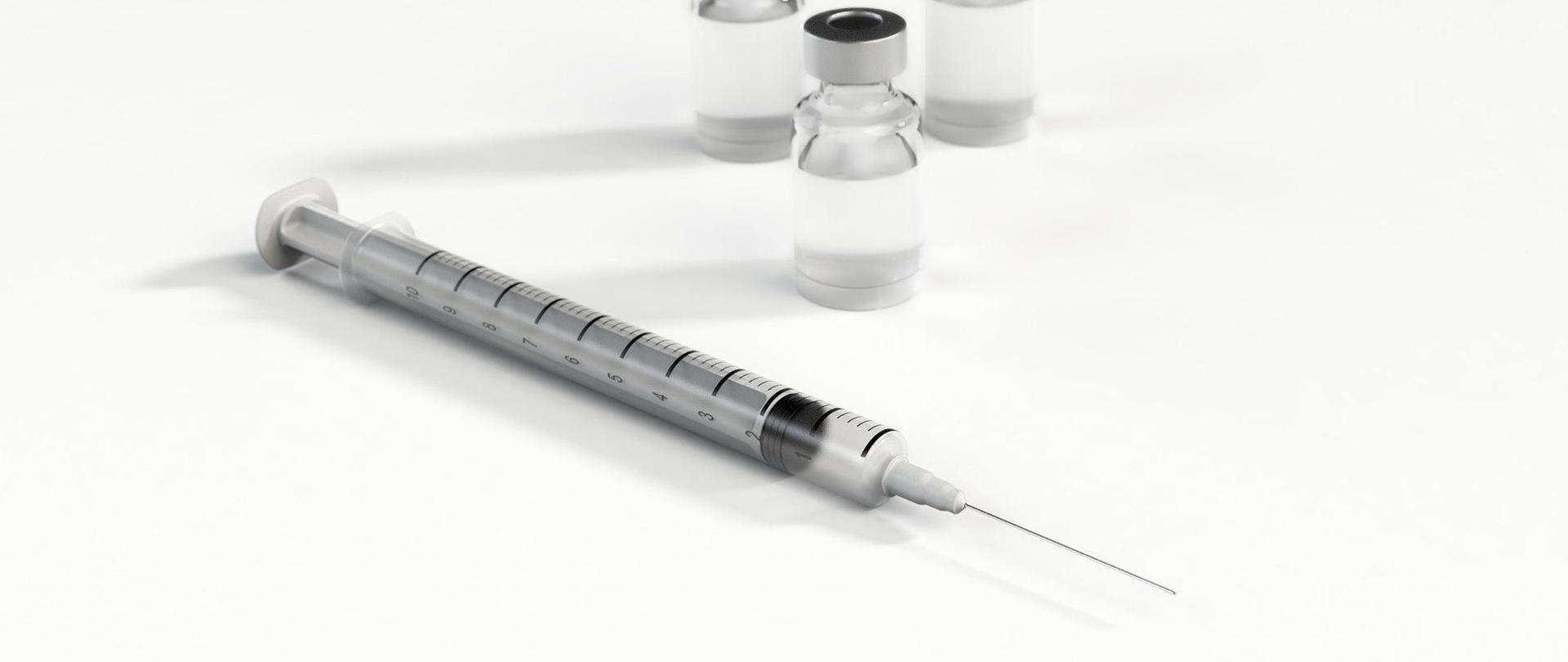 Zdjęcie, na którym znajduje się strzykawka. W tle widoczne są fiolki ze szczepionką.