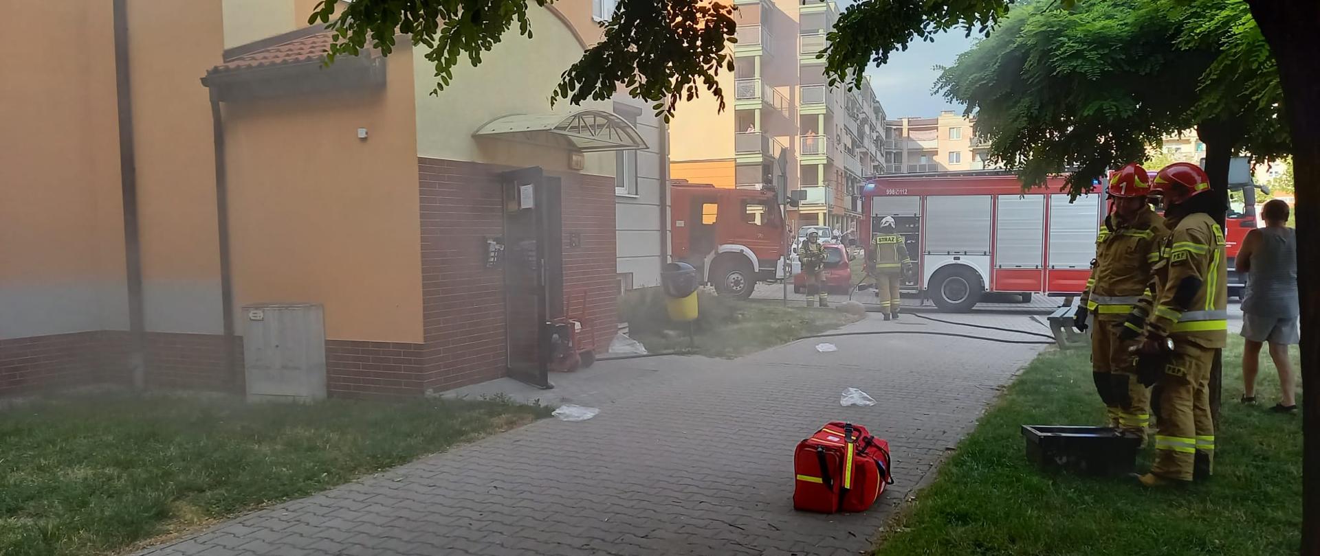 Na zdjęciu widać mur budynku. Przed budynkiem kostka brukowa i zielona trawa. Widać sprzęt straży pożarnej. Zdjęcie zrobione w ciągu dnia. Delikatne zadymienie.