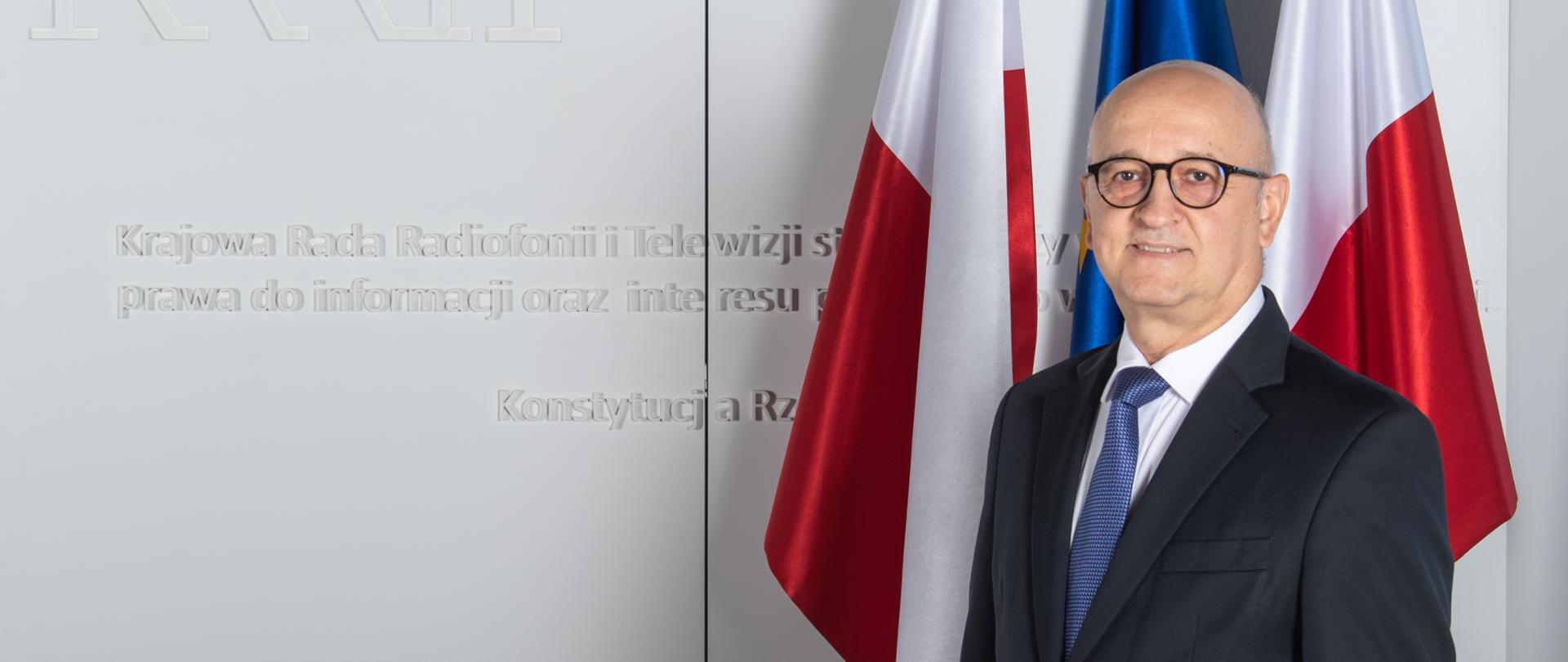 Członek KRRiT Tadeusz Kowalski, za plecami dwie biało-czerwone flagi, pomiędzy nimi niebieska fala Unii Europejskiej 