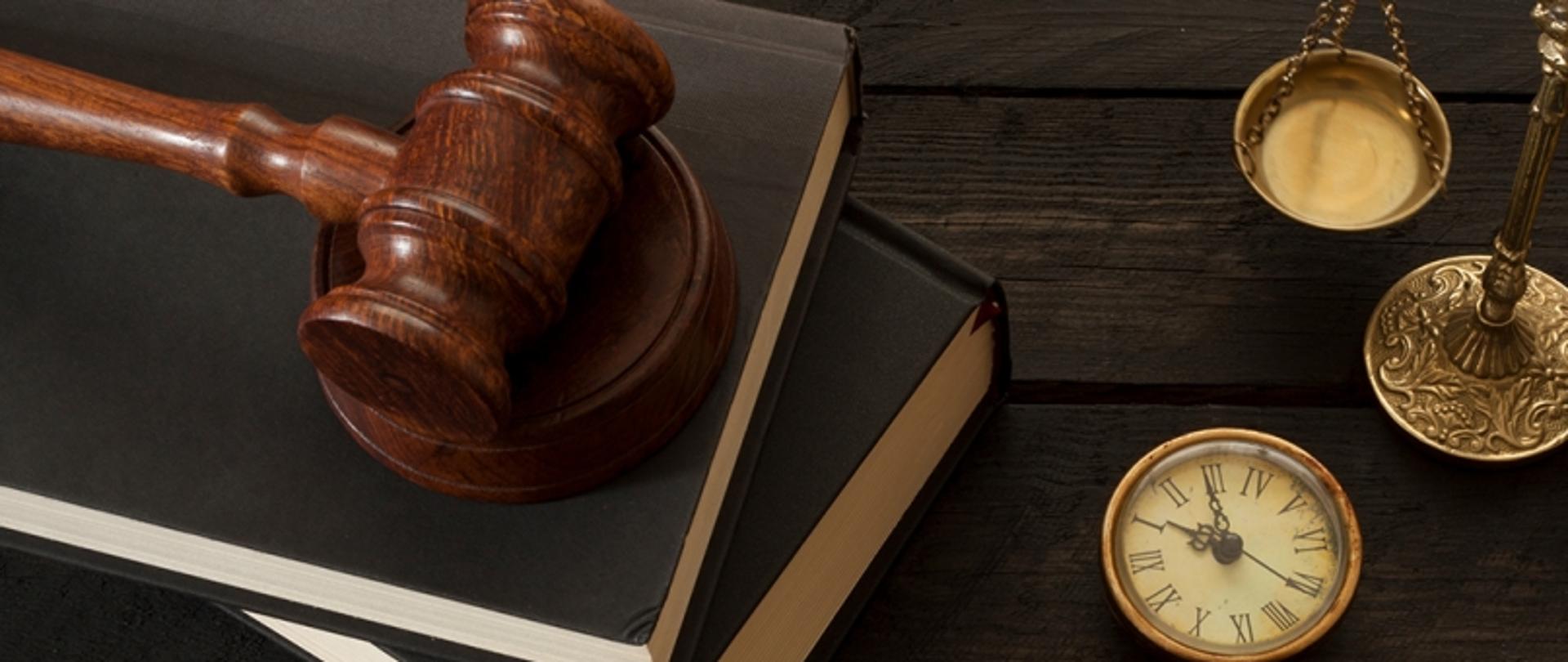 Drewniany młotek sędziowski leżący na książce, obok waga i zegarek na ciemnym drewnianym stole.