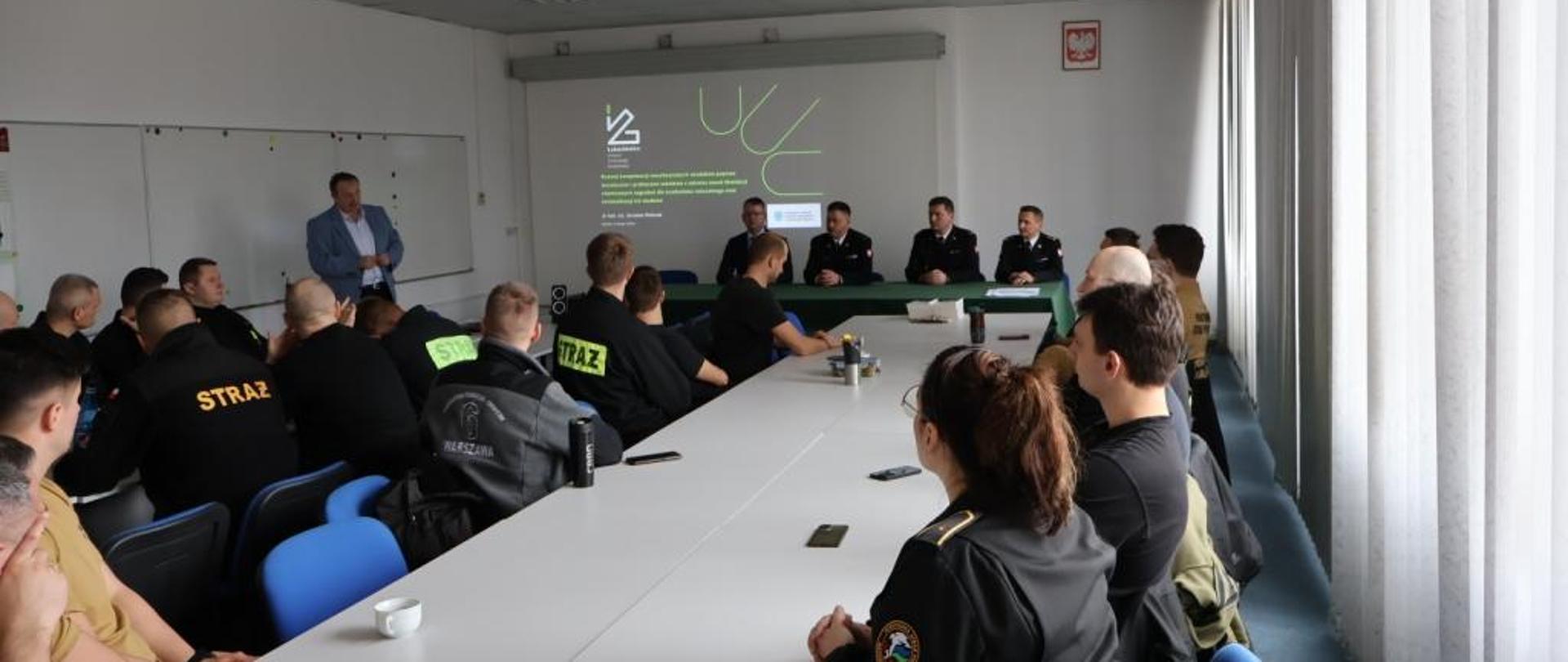 Zdjęcie wykonane w jednym z pomieszczeń szkoleniowych instytutu technologii eksploatacji sieci badawczej łukasiewicz przy ulicy Kazimierza Pułaskiego w Radomiu. Na zdjęciu widać siedzących przy stołach funkcjonariuszy Państwowej Straży Pożarnej. Strażacy ubrani są w ubrania koszarowe oraz dowódczo - sztabowe w czarnym kolorze. W oddali widać osobę wygłaszająca prelekcję. Na ekranie znajdującym się na ścianie pomieszczenia wyświetlana jest prezentacja. 