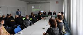 Zdjęcie wykonane w jednym z pomieszczeń szkoleniowych instytutu technologii eksploatacji sieci badawczej łukasiewicz przy ulicy Kazimierza Pułaskiego w Radomiu. Na zdjęciu widać siedzących przy stołach funkcjonariuszy Państwowej Straży Pożarnej. Strażacy ubrani są w ubrania koszarowe oraz dowódczo - sztabowe w czarnym kolorze. W oddali widać osobę wygłaszająca prelekcję. Na ekranie znajdującym się na ścianie pomieszczenia wyświetlana jest prezentacja. 