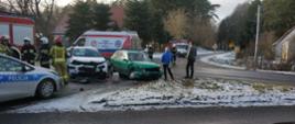 Zdjęcie wykonane w dzień. Zdjęcie przedstawia miejsce zdarzenia. Widać na nim dwa uszkodzone pojazdy, auto policji i auta straży pożarnej oraz karetkę pogotowia. Strażacy oraz służby biorące udział w zdarzeniu wykonują swoje czynności służbowe