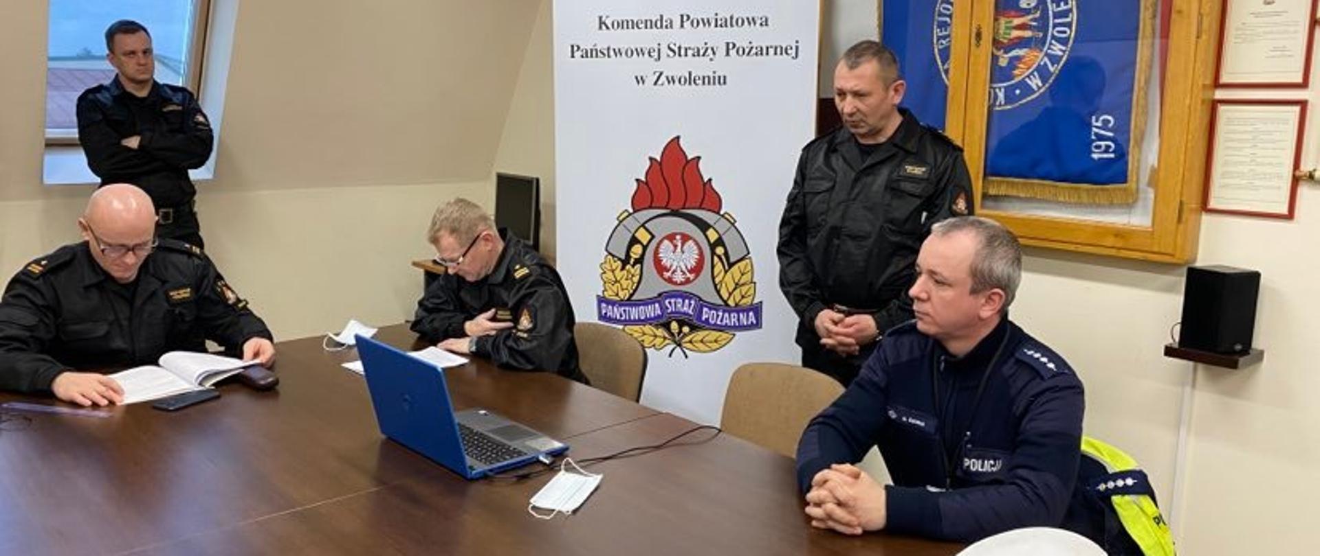 4 osoby na zdjęciu ubrane w ubrania służbowe, w tle rollup Komendy Powiatowej, na stole niebieski laptop