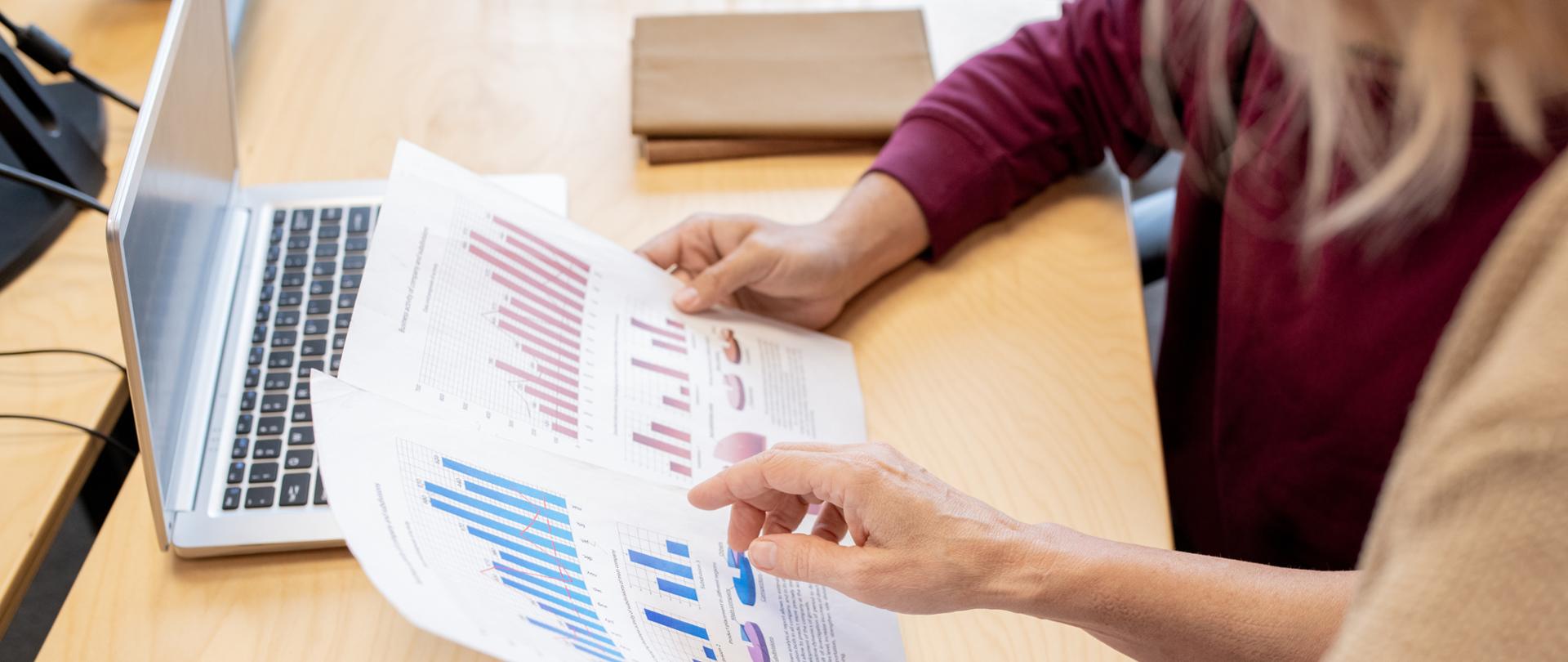 Hand of contemporary female broker pointing at one of financial papers held by colleague while discussing chart and graph by desk